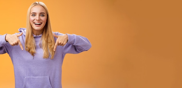 Free photo amused joyful nice blond girlfriend pointing down present cool new product smiling broadly recommending try check out standing orange background happily grinning in hoodie