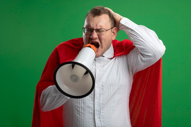 Angry adult slavic superhero man in red cape wearing glasses keeping hand on head looking at side shouting in loud speaker isolated on green wall