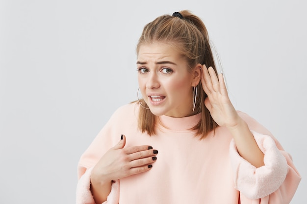 Angry aggressive caucasian female in pink sweater looking with anger and appeal, pointing at herself, holding hand behind her ear like she doesn`t hear what she is told to do, ferusing to listen.