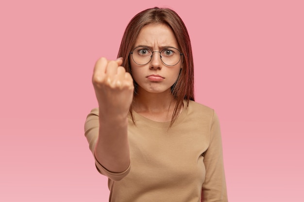 Free photo angry beautiful young woman shows fist , threats someone, being annoyed with noisy neighbours, purses lower lip, dressed in casual clothes