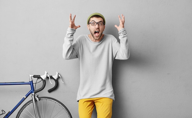 Angry bike instructor screaming with furious expression and rising hands, being angry with his pupils who don`t understand anything. Young cyclist shouting with serious expression, isolated on grey