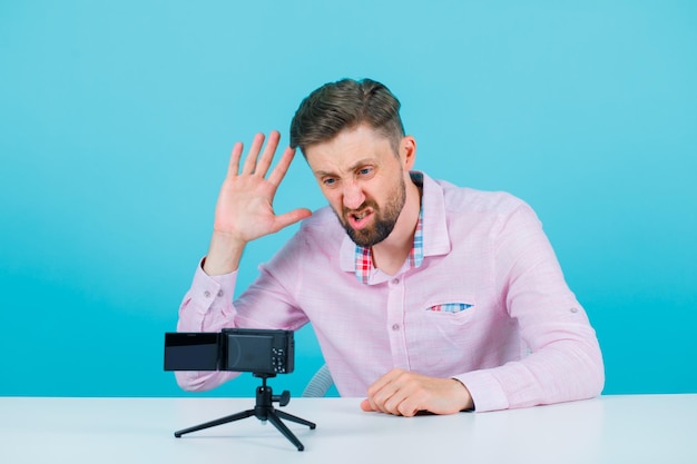 Free photo angry blogger ma is raising up his handful by sitting in front of his mini camera on blue background