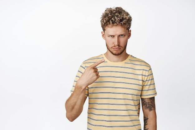 Angry blond guy sulking pointing at himself with upset face expression what about me gesture standing in tshirt over white background