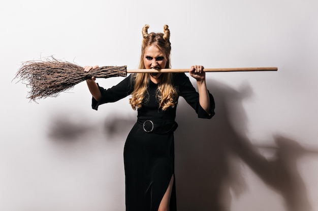 Free photo angry blonde witch standing on white wall. vampire girl posing with broom in halloween.