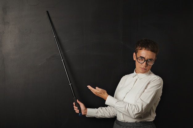 Free Photo angry brunette coach in business suit with bad emotion and shows on black chalk board behind her with folding pointer and other hand.