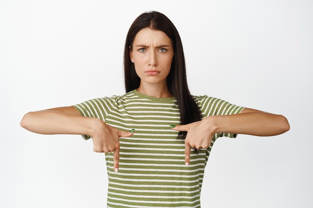 Free Photo angry brunette woman frowning and sulking pointing fingers up at smth bad standing in tshirt over white background