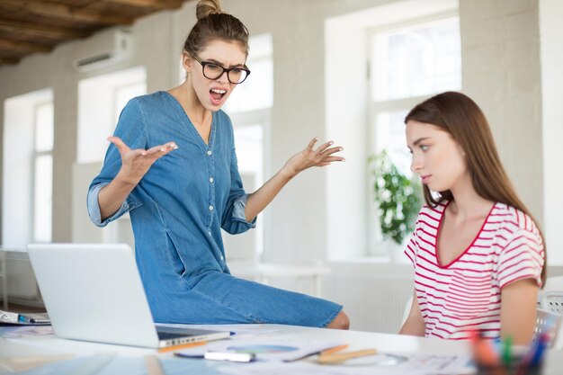 Angry business woman in eyeglasses leaning on desk while screaming on employee in modern office