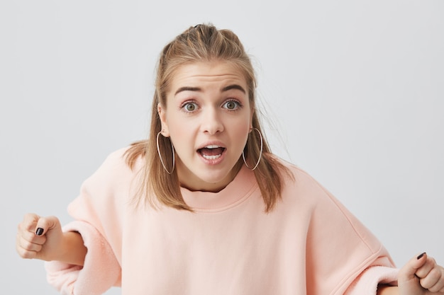 Angry caucasian young female with healthy clean skin wearing pink sweater posing isolated , popping her eyes, ready to argue. Negative emotions, dissatisfaction, aggression.