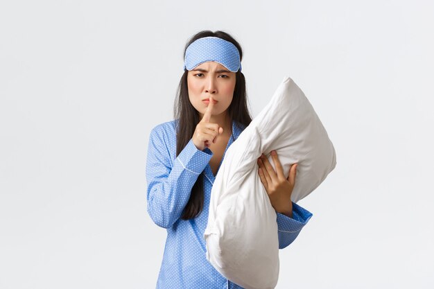 Angry displeased asian girl in sleeping mask and pyjamas, holding pillow and frownign disturbed, shushing, say shhh as woke-up from loud noise, asking people keep quiet at night, white background.