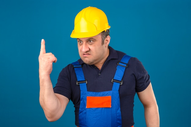 Angry in furious middle aged man wearing construction uniform and safety helmet pointing finger up displeased and frustrated over isolated blue wall