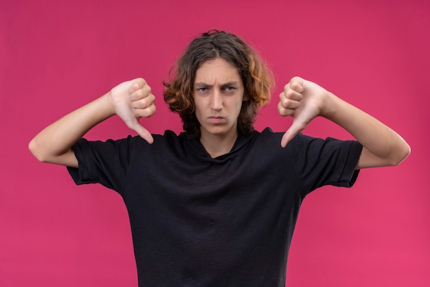 Free photo angry guy with long hair in black t-shirt shows thumb down on pink wall