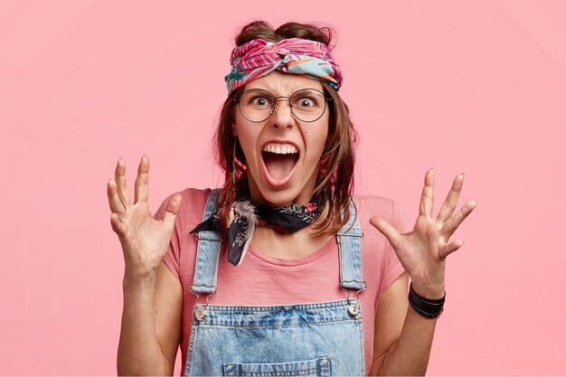 Angry hippy woman shouts with irritation and gestures in annoyance, has nervous facial expression, recieves bad news, stands over pink wallimpatient