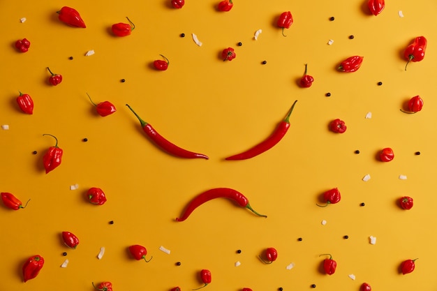 Free photo angry human face made of red chili pepper, other peppers arranged around on yellow background. spicy vegetable that can trigger burning sensation and cause health problems, has own distinct taste