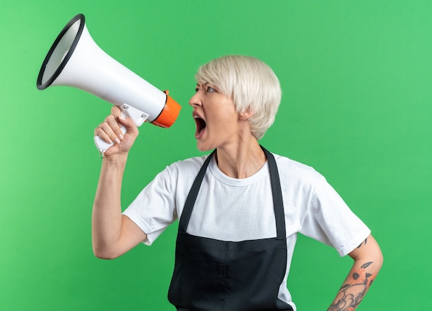 Angry looking at side young beautiful female barber in uniform speaks on loudspeaker isolated on green wall