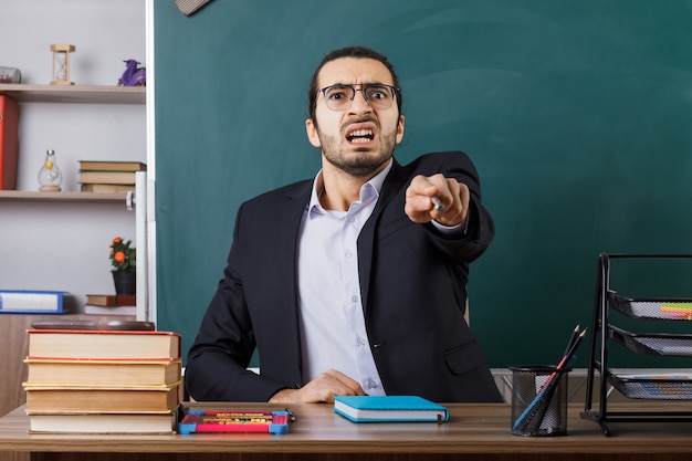 Free Photo angry male teacher wearing glasses points with pointer stick sitting at table with school tools in classroom