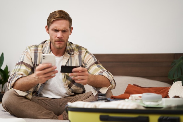 Free photo angry man holds credit card and mobile phone sits next to clothes in suitcase has problems with