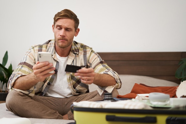 Free photo angry man holds credit card and mobile phone sits next to clothes in suitcase has problems with