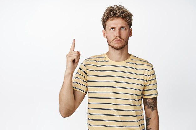 Angry and moody blond man pointing finger up looking with upset frowning face at advertisement standing annoyed against white background