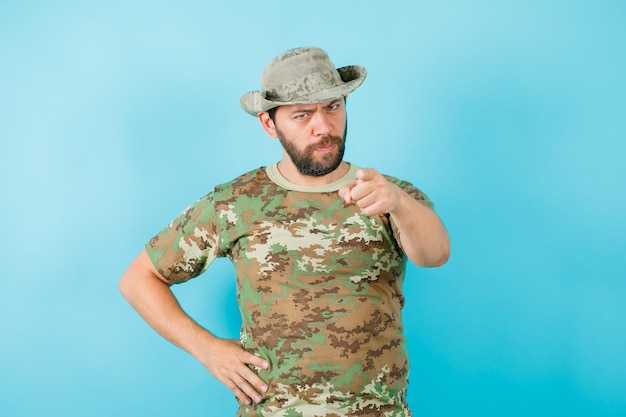 Free Photo angry officer is pointing camera with forefinger and putting other hand on waist on blue background