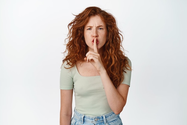 Free Photo angry redhead girl shushing press finger to lips and hush showing taboo gesture standing in tshirt against white background