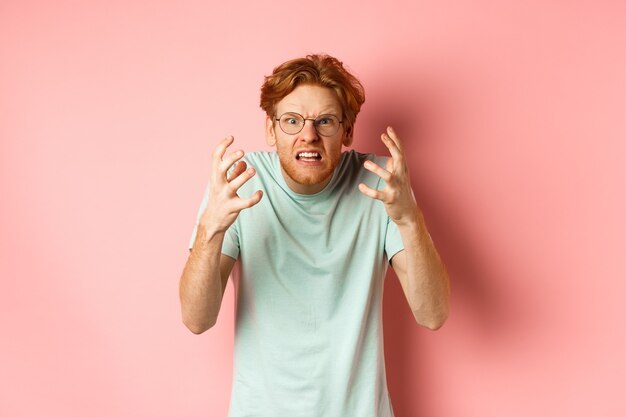 Angry redhead guy in glasses shouting, frowning and shaking hands with frustrated and outraged face, standing over pink background.