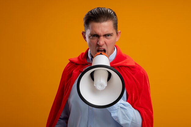 Angry super hero businessman in red cape shouting to megaphone with aggressive expression standing over orange wall