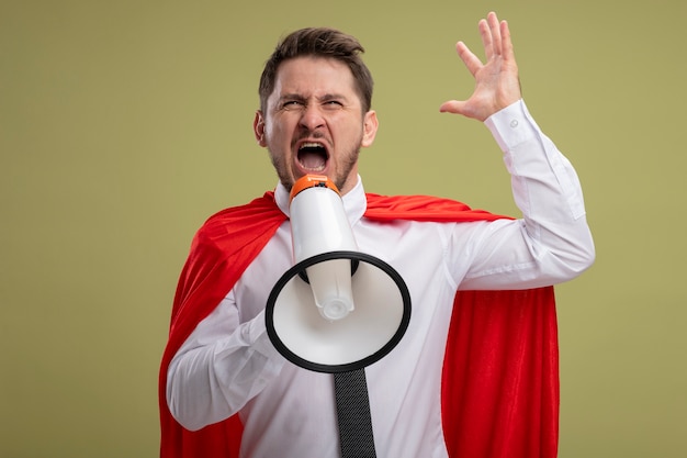 Free photo angry super hero businessman in red cape shouting to megaphone with aggressive expression with raised arm standing over green background