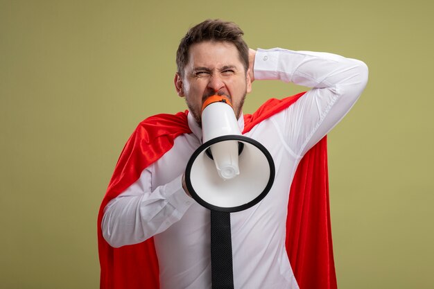 Angry super hero businessman in red cape shouting to megaphone with aggressive expression with raised hand standing over green background