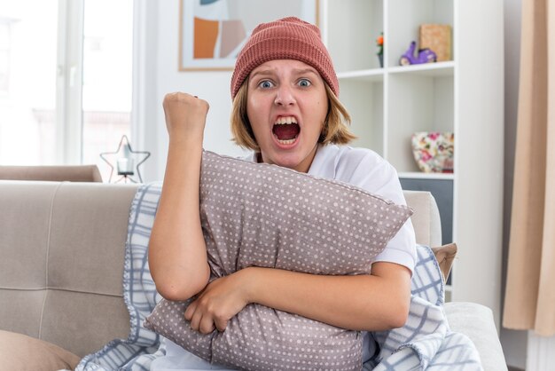 Free photo angry unhealthy young woman in warm hat with blanket looking unwell suffering from cold and flu holding pillow clenching fist shouting with aggressive expression sitting on couch in light living room