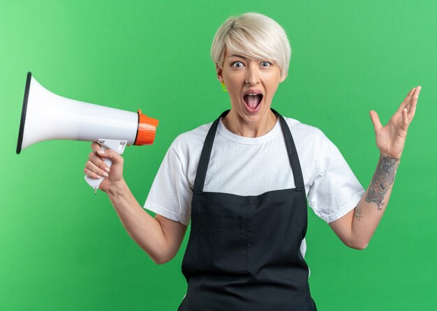 Angry young beautiful female barber in uniform holding loudspeaker isolated on green wall