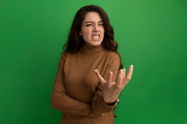 Angry young beautiful girl holding out hand at camera isolated on green wall