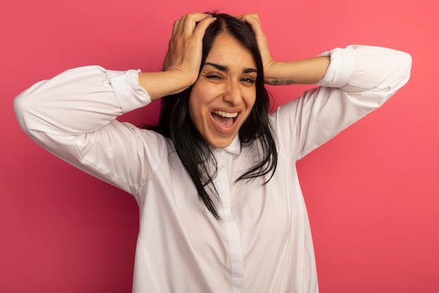 Angry young beautiful girl wearing white t-shirt grabbed head isolated on pink