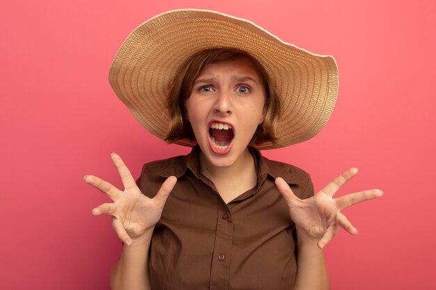 Angry young blonde girl wearing beach hat looking  showing empty hands isolated on wall