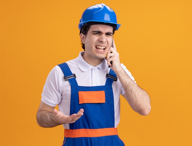 Angry young builder man in construction uniform and safety helmet talking on mobile phone standing over orange wall