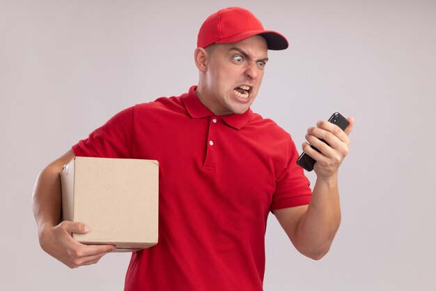 Angry young delivery man wearing uniform with cap holding box and looking at phone in his hand isolated on white wall