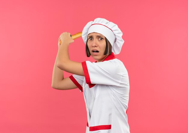 Angry young female cook in chef uniform holding bread stick and getting ready to beat someone isolated on pink  with copy space