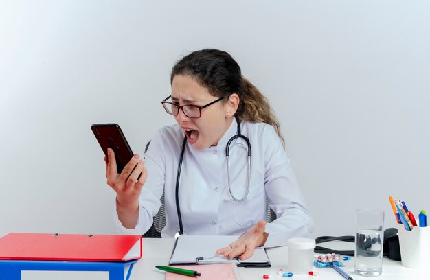 Angry young female doctor wearing medical robe and stethoscope and glasses sitting at desk with medical tools holding looking at mobile phone isolated