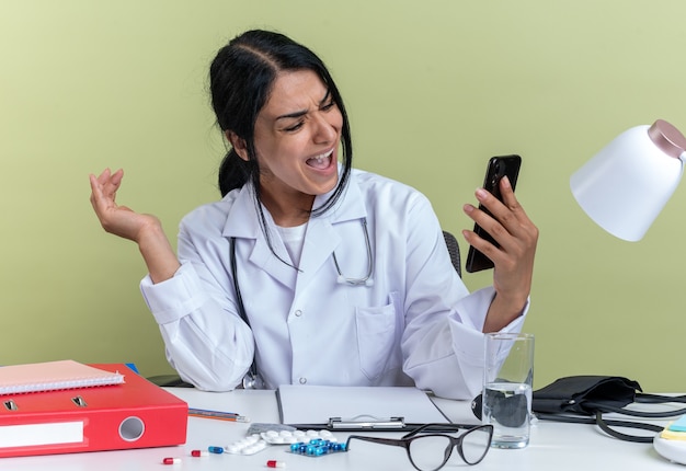 Free Photo angry young female doctor wearing medical robe with stethoscope sits at desk with medical tools holding and looking at phone isolated on olive green wall