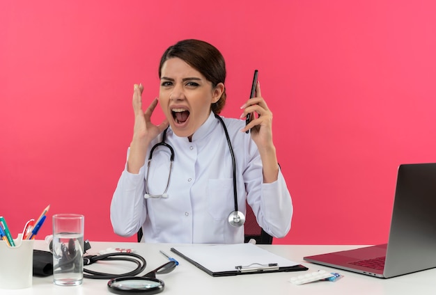 Free Photo angry young female doctor wearing medical robe with stethoscope sitting at desk work on computer with medical tools speakes on phone on isolated pink wall with copy space