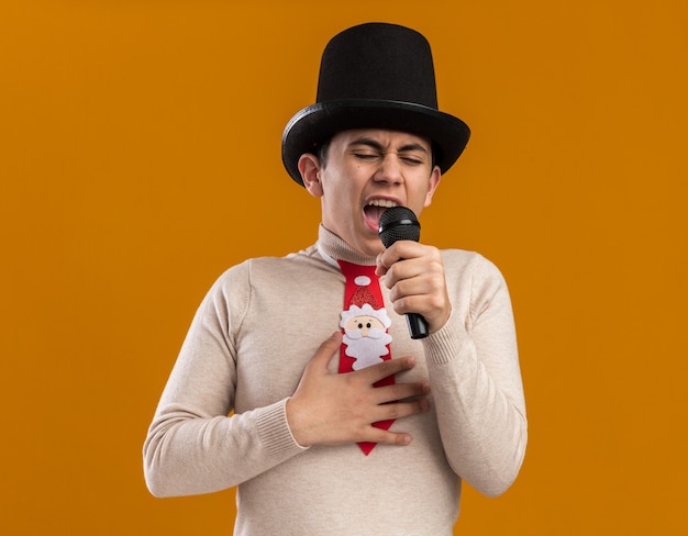 Angry young guy wearing hat with christmas tie speaks on microphone isolated on yellow wall