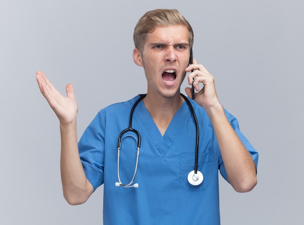 Free Photo angry young male doctor wearing doctor uniform with stethoscope speaks on phone spreading hand isolated on white wall