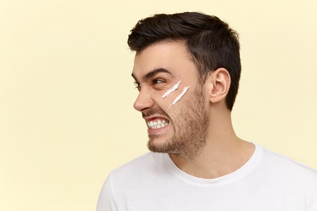 Angry young man doing morning routine with lotion cream on his face