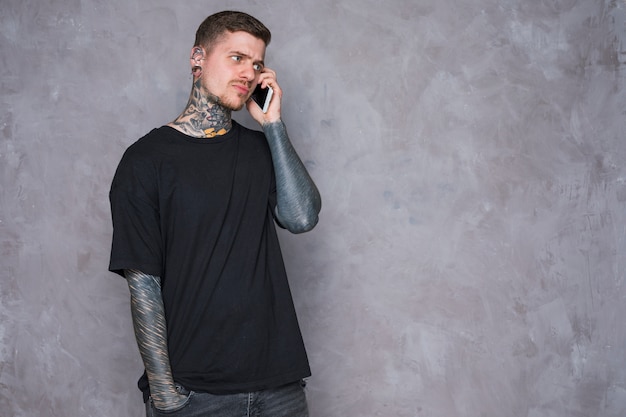 An angry young man with tattooed on his hand talking on mobile phone against grey wall