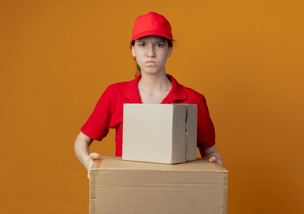 Free Photo angry young pretty delivery girl in red uniform and cap holding carton boxes looking at camera isolated on orange background with copy space