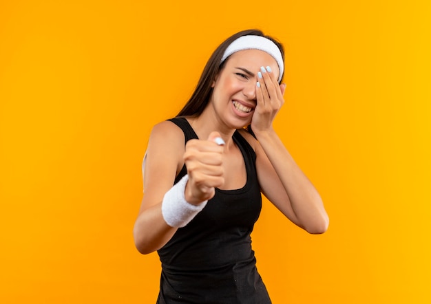 Angry young pretty sporty girl wearing headband and wristband stretching out her fist  and putting her hand on eye suffering from pain isolated on orange wall with copy space