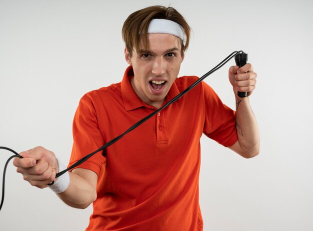 Angry young sporty guy wearing headband with wristband stretching jump rope isolated on white wall