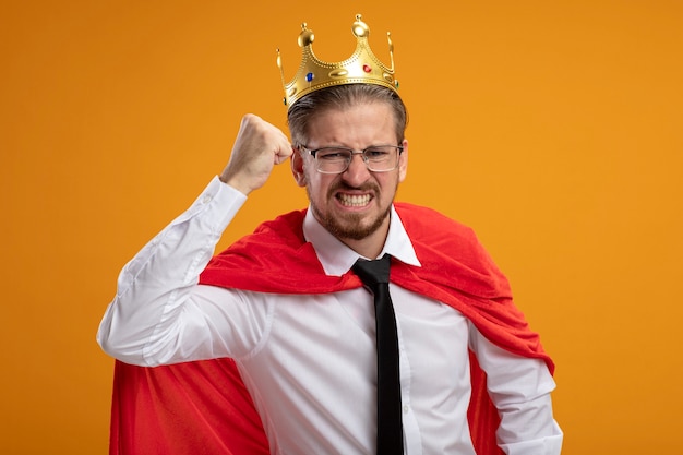 Free photo angry young superhero guy wearing tie and crown with glasses raising fist isolated on orange