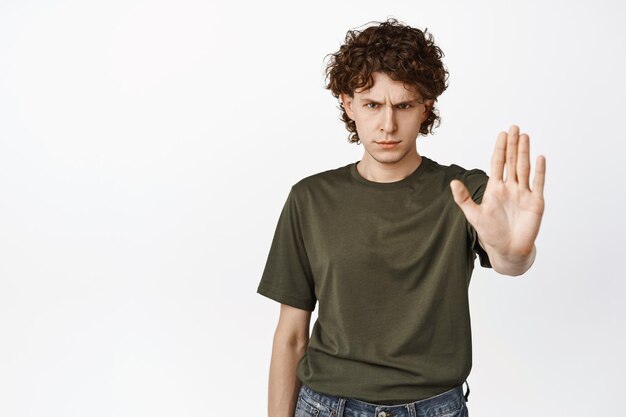 Angry young teenage man stretch out hand showing stop refusal gesture prohibit smth standing serious against white background