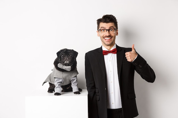 Animals, party and celebration concept. Handsome young man in suit and cute black pug in costume staring at camera, owner showing thumb up in approval and praise, white background