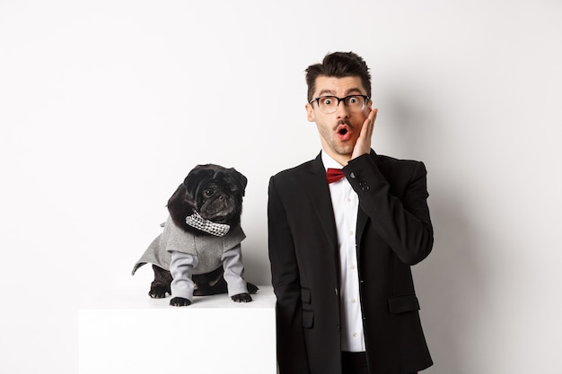 Animals, party and celebration concept. Shocked handsome man in formal suit and cute dog in costume, staring at camera amazed, standing over white background.